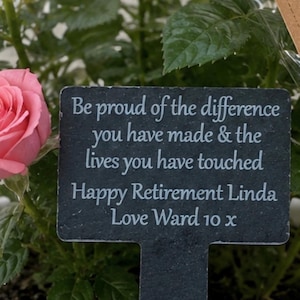 May include: A rectangular black slate plant marker with white engraved text that reads: Be proud of the difference you have made & the lives you have touched. Happy Retirement Linda. Love Ward 10 x. The marker is placed in a white pot with a pink rose.
