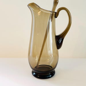 May include: A brown glass jug with a matching glass stirrer. The jug has a wide base and a rounded body with a flared lip. The handle is attached to the side of the jug.