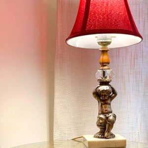 May include: A vintage table lamp with a red lampshade and a decorative base. The base features a cherub figure holding up a crystal accent. The lamp sits on a square marble base, adding a touch of elegance to any room.