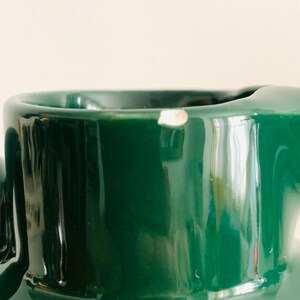 May include: Close-up of a glossy, dark green ceramic mug. The mug has a smooth, reflective surface with a small chip near the rim. The interior is not visible. The background is a soft, off-white color.