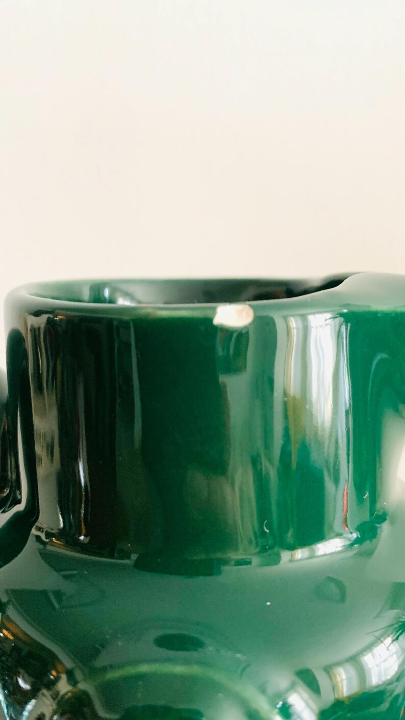 May include: Close-up of a glossy, dark green ceramic mug. The mug has a smooth, reflective surface with a small chip near the rim. The interior is not visible. The background is a soft, off-white color.