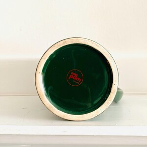 May include: Bottom view of a green ceramic mug with a dark green interior. The base features a red circular logo with white text. The rim is a cream color. The mug is on a white surface.