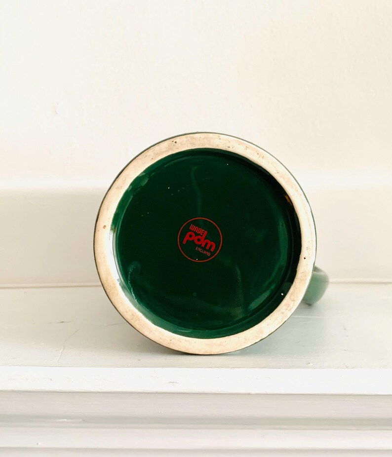 May include: Bottom view of a green ceramic mug with a dark green interior. The base features a red circular logo with white text. The rim is a cream color. The mug is on a white surface.
