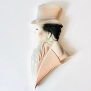 May include: A ceramic wall hanging depicting a person in profile, wearing a top hat and formal attire. The figure has black hair, rosy cheeks, and is dressed in shades of pink and white. The piece is likely vintage.