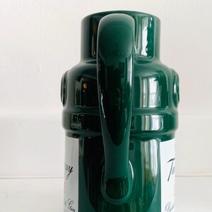 May include: A dark green ceramic pitcher with a large handle. The pitcher has a cylindrical body with a rounded top and a label with text. The pitcher is on a white surface.