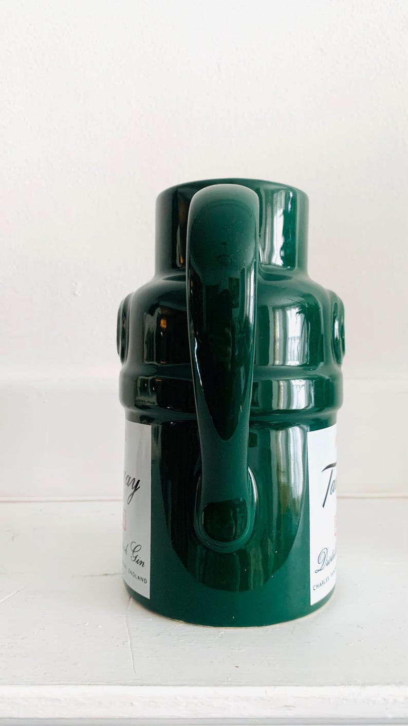 May include: A dark green ceramic pitcher with a large handle. The pitcher has a cylindrical body with a rounded top and a label with text. The pitcher is on a white surface.