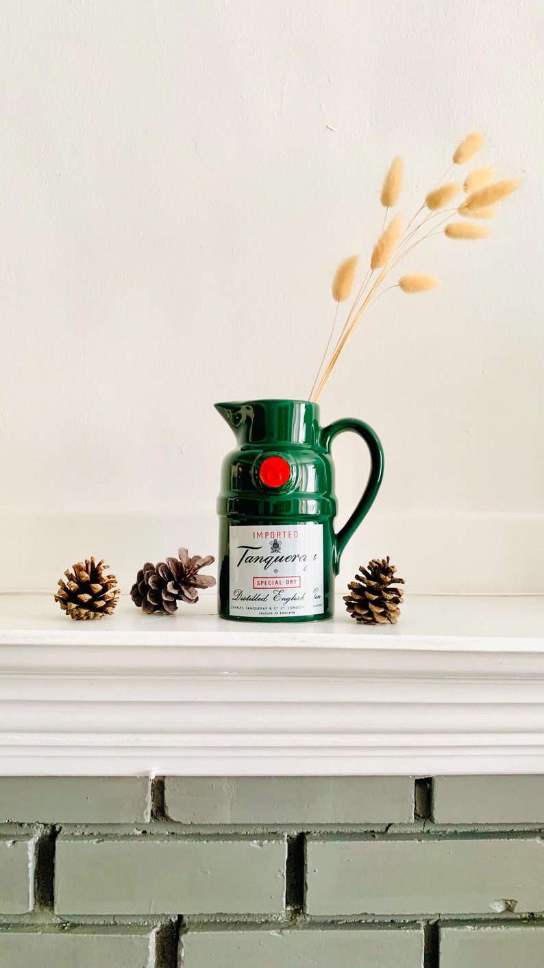 May include: A green ceramic pitcher with a red accent and the text "Tanqueray Special Dry" is displayed with dried flowers and pinecones. The pitcher is a decorative item, possibly for a bar or kitchen.