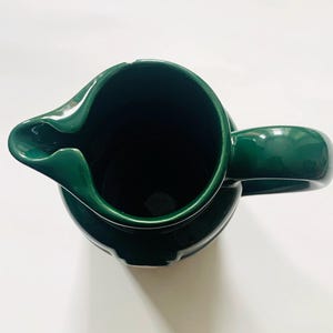 May include: A top-down view of a dark green ceramic pitcher with a curved spout and a rounded handle. The pitcher has a glossy finish and a wide opening. The interior of the pitcher is dark.