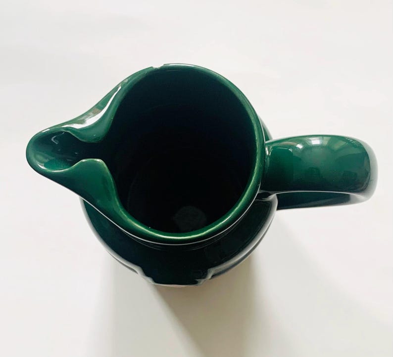 May include: A top-down view of a dark green ceramic pitcher with a curved spout and a rounded handle. The pitcher has a glossy finish and a wide opening. The interior of the pitcher is dark.
