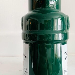 May include: A dark green ceramic bottle with a glossy finish. The bottle has a unique shape with a rounded body and a narrow neck. White labels with text are visible on the bottle. The bottle is likely for spirits or beverages.