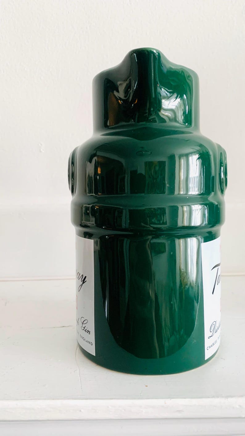 May include: A dark green ceramic bottle with a glossy finish. The bottle has a unique shape with a rounded body and a narrow neck. White labels with text are visible on the bottle. The bottle is likely for spirits or beverages.