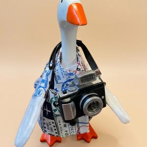 May include: A white goose figurine with an orange beak and feet, wearing a white shirt patterned with blue and black travel stamps and passport markings. The goose has a black camera with a silver lens and a flash unit hanging around its neck.