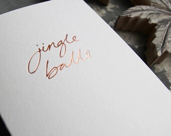 Könnte beinhalten: Eine weiße Karte mit dem Schriftzug "jingle balls" in kupferfarbener Folie.