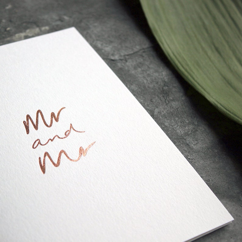 May include: A white card with the words "Mr and Mr" written in copper foil.