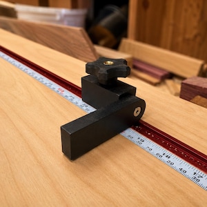 Puede incluir: Un tope de guía para carpintería de metal negro con una perilla de ajuste en forma de estrella, colocado sobre un riel de aluminio rojo con una escala de medición blanca en unidades métricas, sobre una superficie de madera clara.
