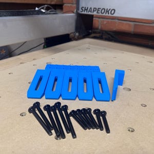May include: A collection of blue, rectangular clamps with elongated openings, and a set of black screws. The clamps are arranged on a light-colored wooden surface, likely for a CNC machine.