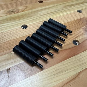 May include: A collection of eight black knobs with cylindrical handles and thin, protruding stems. The knobs are arranged diagonally on a light-colored wooden surface, showcasing their design and potential use in woodworking or furniture.