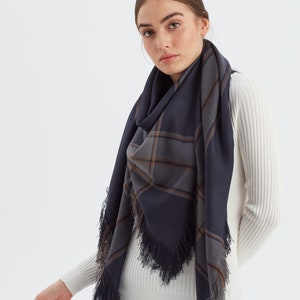 May include: A navy blue and grey plaid scarf with fringe trim. The scarf is draped around the neck of a person wearing a white sweater and black trousers.