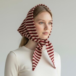 Puede incluir: Un pañuelo a rayas rojas y crema atado alrededor de la cabeza y el cuello. El pañuelo está hecho de un material suave y presenta un patrón de rayas clásico. El cubrecabezas es un accesorio elegante.