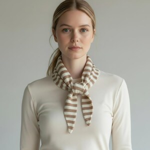 May include: A beige and brown striped scarf tied in a knot. The scarf is made of a soft, knitted material. The person is wearing a cream-coloured long-sleeved top.