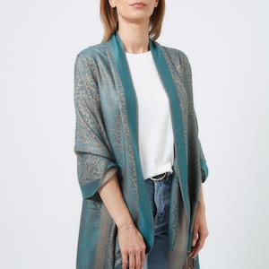 May include: A teal and brown patterned shawl with fringe. The shawl is draped over a person's shoulders and is worn with a white t-shirt and blue jeans.
