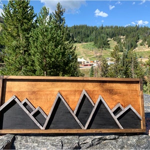 May include: A wooden wall art piece featuring a mountain range silhouette. The mountains are made of black wood with silver accents and are set against a light wood background.