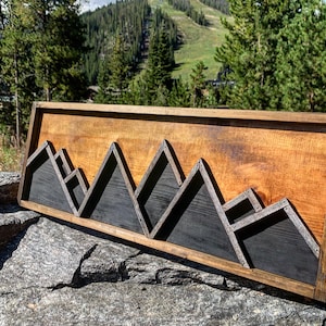 May include: A wooden wall art piece featuring a mountain range silhouette. The mountains are made of dark stained wood and are set against a light wood background. The piece is framed in a dark wood frame.