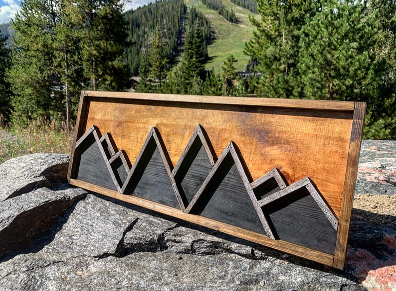 May include: A wooden wall art piece featuring a mountain range silhouette. The mountains are made of dark stained wood and are set against a light wood background. The piece is framed in a dark wood frame.