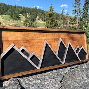 May include: A wooden wall art piece depicting a mountain range with a black and gray color scheme. The mountains are framed in a dark wood frame.