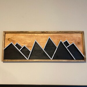 May include: A wooden wall art piece featuring a mountain range silhouette with a black and white color scheme. The mountains are painted black with white outlines. The wood background is a light brown color.