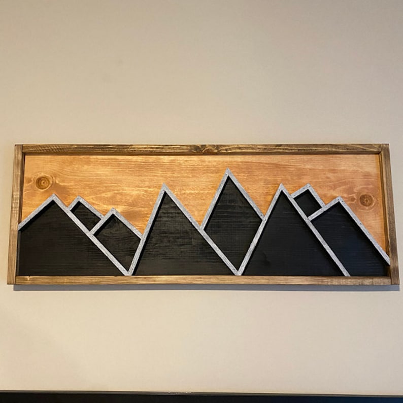 May include: A wooden wall art piece featuring a mountain range silhouette with a black and white color scheme. The mountains are painted black with white outlines. The wood background is a light brown color.