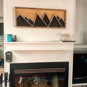 May include: A wooden wall art piece with a black mountain range design. The art is mounted above a white fireplace mantel with a black fireplace.