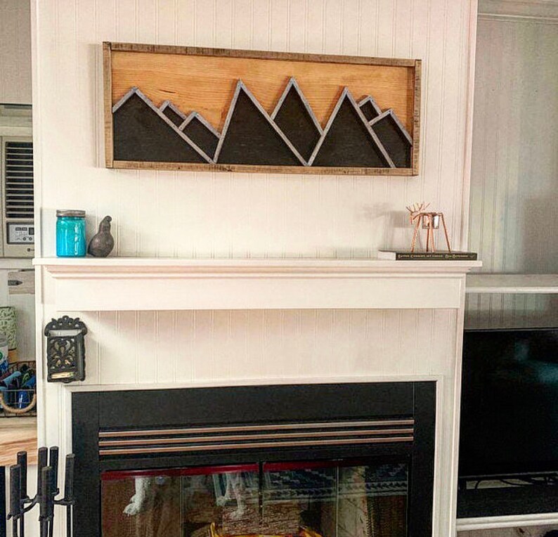 May include: A wooden wall art piece with a black mountain range design. The art is mounted above a white fireplace mantel with a black fireplace.
