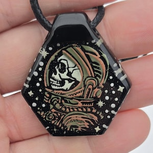 May include: Black, hexagonal pendant necklace featuring a skull in an astronaut helmet design. The pendant has a black background with white star accents. The necklace has a black cord.