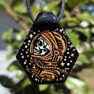 May include: A black hexagonal pendant necklace with a gold astronaut skull design on a black background with white stars.