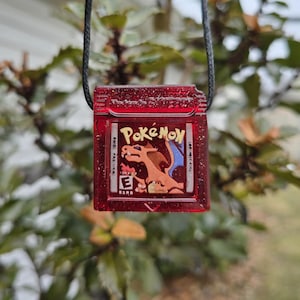 May include: A red Pokémon cartridge pendant on a black cord. The cartridge displays the Pokémon logo and a Charizard graphic. This pendant is a nostalgic item for fans of the popular video game series.