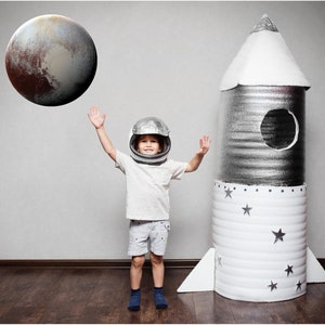 May include: A young child dressed as an astronaut, wearing a white helmet and a white shirt with grey shorts, raises their arms in the air in front of a large cardboard rocket ship. A large, round, brown and grey planet is in the background.