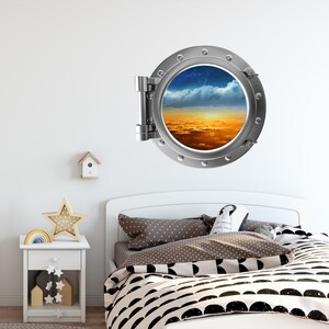 May include: A silver porthole wall decal with a view of a sunset over clouds. The decal is mounted on a white wall above a white bed with black and white bedding.