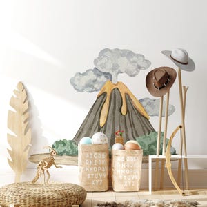 May include: A wall decal featuring a gray and yellow volcano with smoke clouds, positioned behind two tan fabric storage bins printed with the alphabet. A wooden dinosaur skeleton, a wooden bow, and two hats on a stand are also present.