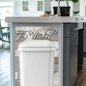 May include: A white rectangular trash can with a lid, set against a backdrop of weathered wood-look paneling. The word "TRASH" is displayed in elegant white script above the can. The kitchen setting includes a countertop and cabinets.