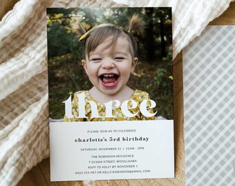 Op de afbeelding: Een verjaardagsuitnodiging met een foto van een jong meisje dat lacht. De uitnodiging zegt "three" in grote, kleurrijke letters en bevat de tekst "Please join us to celebrate Charlotte's 3rd birthday." De uitnodiging bevat ook de datum, tijd en locatie van het feest.
