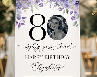 May include: A white sign with purple flowers and a photo of a woman. The sign says "80 eighty years loved Happy Birthday Elizabeth!"