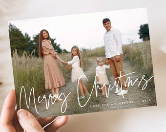 May include: A Christmas card featuring a family of four standing on a path in a field. The parents are holding hands with their two daughters. The card has the text "Merry Christmas" written in white cursive script and "Love the Andersons | 2023" written in white text below the cursive script.