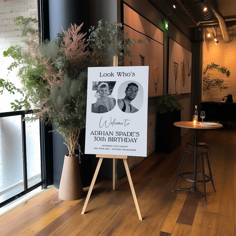 Pu&ograve; includere: Un cartello bianco con testo nero e una foto di due persone. Il cartello recita "Look Who's", "Welcome to", "Adrian Spade's 30th Birthday", "Saturday 5th August", "Cru Wine Bar - Kitchen Melbourne".