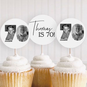May include: Three white circular cupcake toppers on wooden sticks. Two toppers feature the number 70 with black-and-white photos of a person at different ages. The center topper displays the black text Thomas IS 70! in a mix of script and sans-serif fonts.