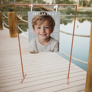 K&ouml;nnte beinhalten: Ein kupferfarbener Metallrahmen h&auml;lt ein Poster mit dem Text "FINLAY IS 5". Das Poster zeigt ein l&auml;chelndes Kind und zus&auml;tzlichen Text. Der Rahmen befindet sich auf einem Holzsteg in der N&auml;he von Wasser.