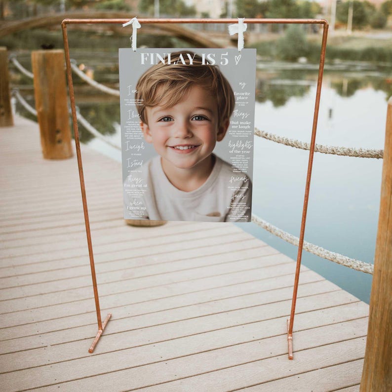 K&ouml;nnte beinhalten: Ein kupferfarbener Metallrahmen h&auml;lt ein Poster mit dem Text "FINLAY IS 5". Das Poster zeigt ein l&auml;chelndes Kind und zus&auml;tzlichen Text. Der Rahmen befindet sich auf einem Holzsteg in der N&auml;he von Wasser.