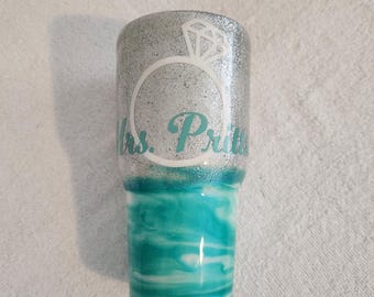 May include: A silver glitter tumbler with a turquoise and white marbled design. The top features a white diamond ring graphic and the words "Mrs. Prit". A unique gift for a bride.
