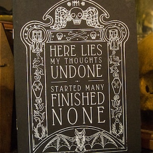 May include: Black and white illustration of a tombstone with the text "Here lies my thoughts undone... Started many finished none". The tombstone is decorated with skulls, crossbones, and a bat.
