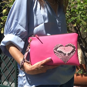 Op de afbeelding: Een roze leren clutch met een hartvormige slangenvellen patch. De clutch heeft een gouden rits en een gouden kettingriem.